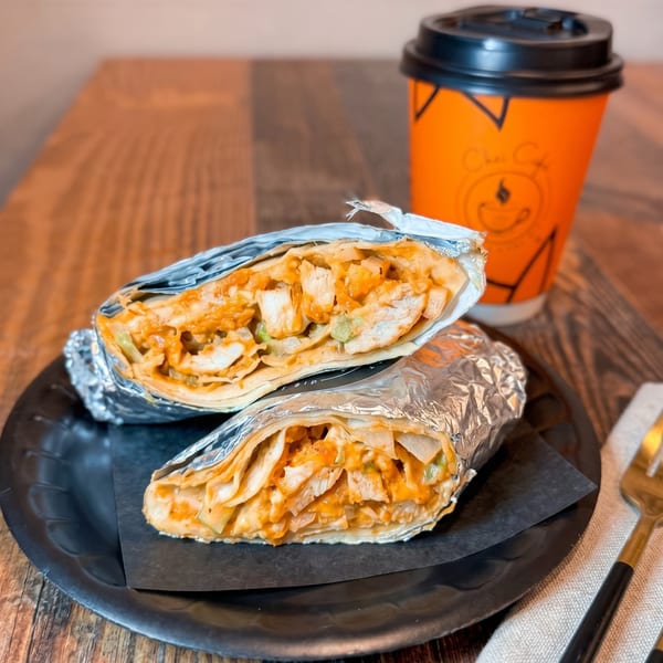 Two foil-wrapped chicken wraps on a black plate with an orange Chai Cafe cup in the background.